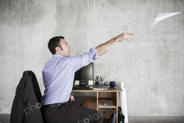 Caucasian businessman throwing a paper airplane while sitting at his desk in a raw…