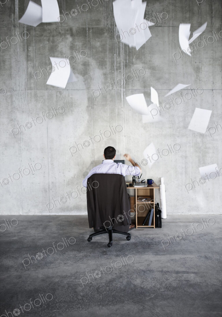 Caucasian businessman throwing papers in air while sitting at his desk in a large raw…
