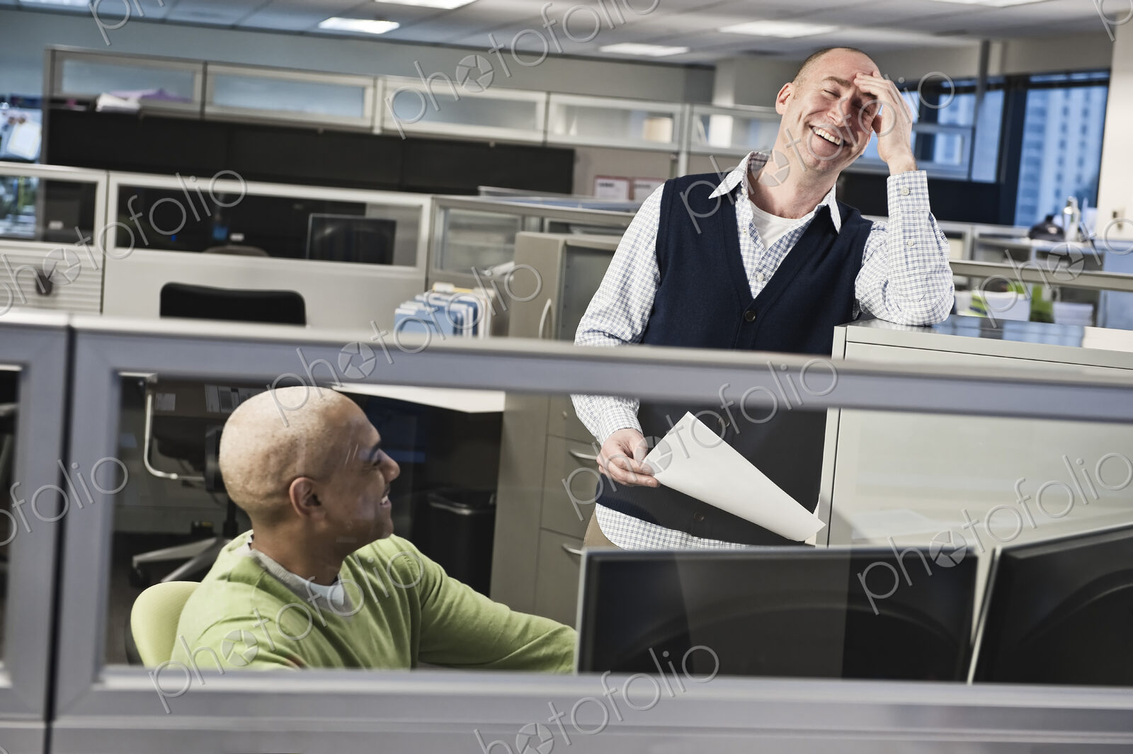 Caucasian male and black male talking and smiling in a cubicle office setting.