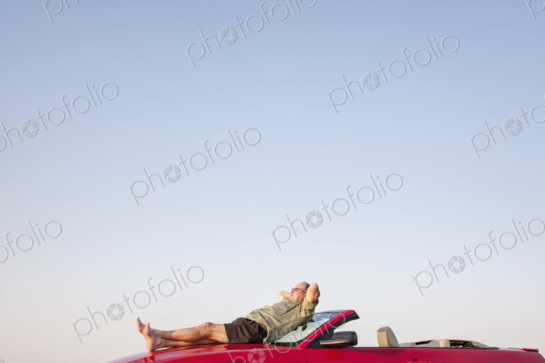 Caucasian male relaxing on the hood of his convertible sports car.