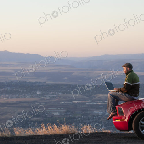 Caucasian male sitting on the hood of his car working on a lap top computer at a rest…