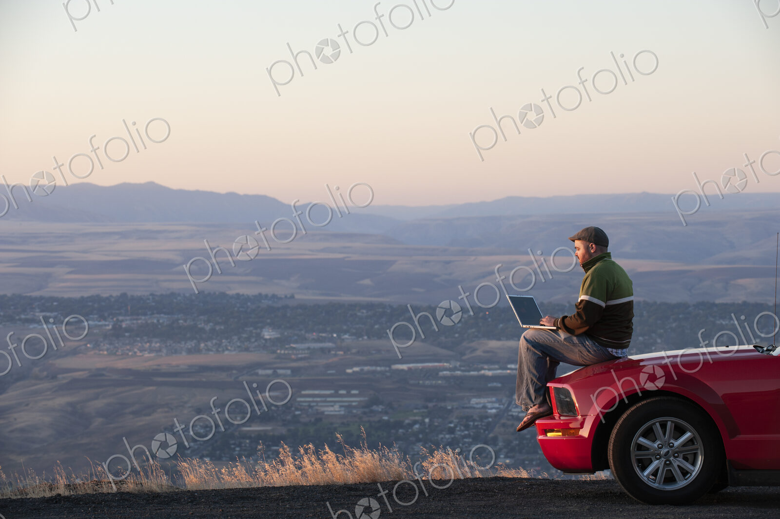 Caucasian male sitting on the hood of his car working on a lap top computer at a rest…