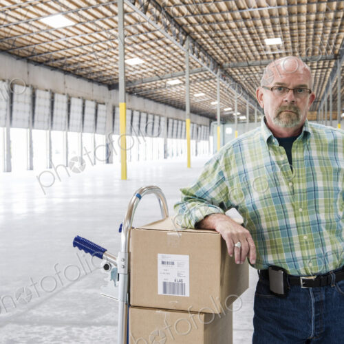 Caucasian male standing next to hand truck in front of loading dock doors inside of a… Caucasian male standing next to hand truck in front of loading dock doors inside of a…
