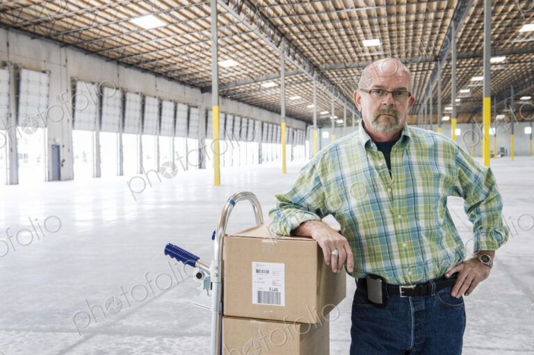 Caucasian male standing next to hand truck in front of loading dock doors inside of a…