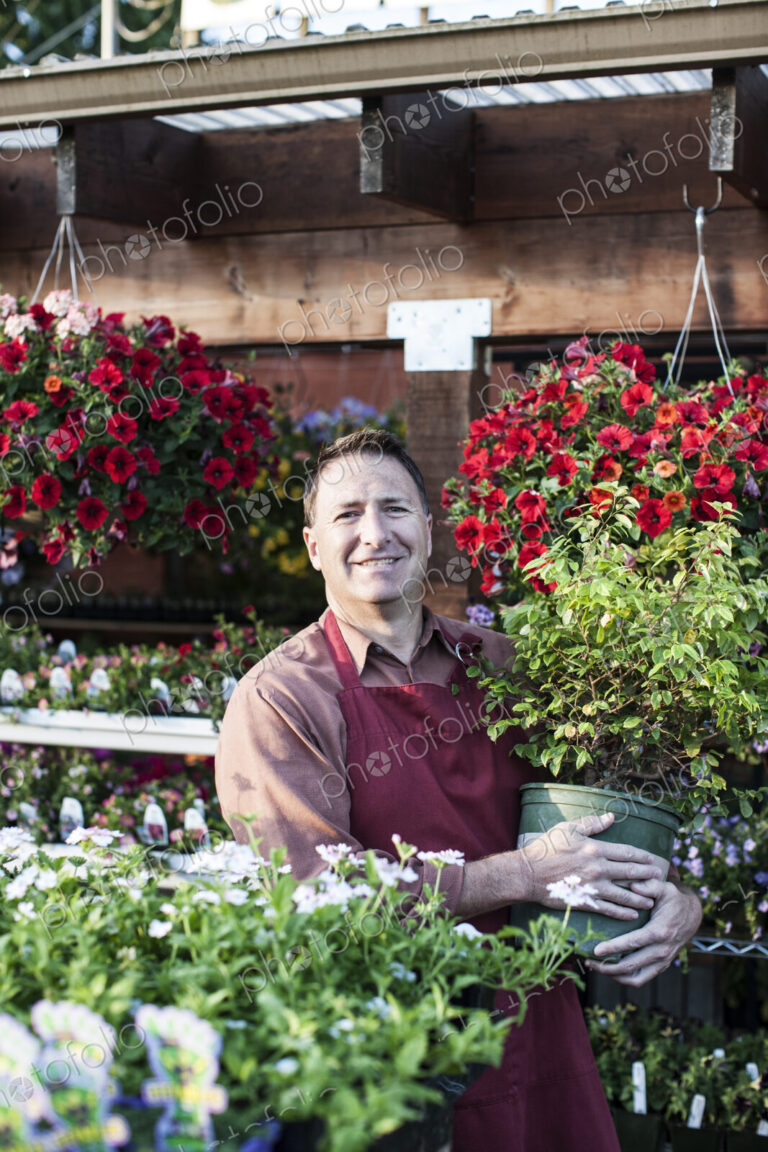 Caucasian man, a nursery owner standing holding a large plant in a pot, by displays of…