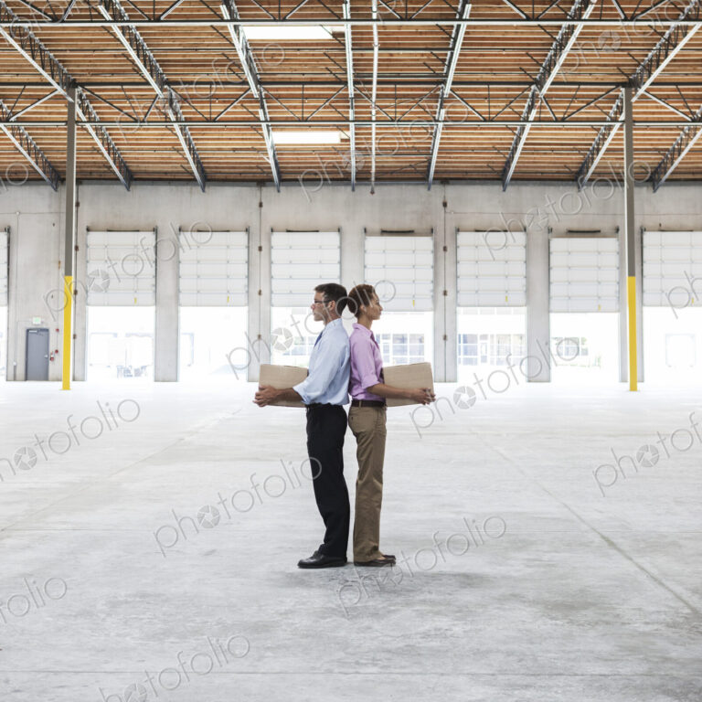 Caucasian man and black female holding cardboard boxes in middle of empty warehouse…