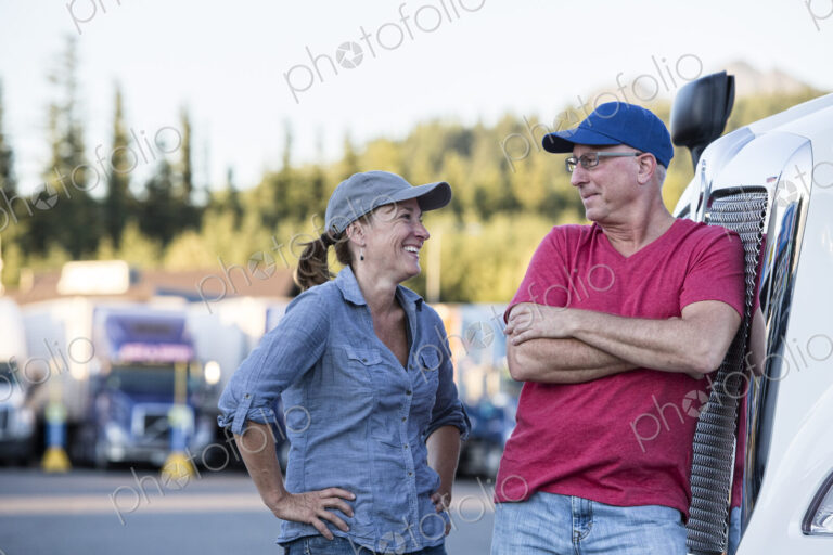 Caucasian man and woman driving team talking in front of a large truck at a truck stop.
