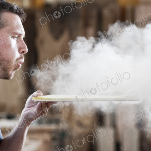 Caucasian man factory worker blowing sanding dust off a wooden piece in a woodworking…