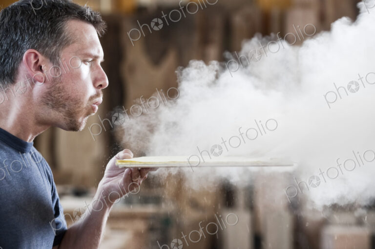 Caucasian man factory worker blowing sanding dust off a wooden piece in a woodworking…