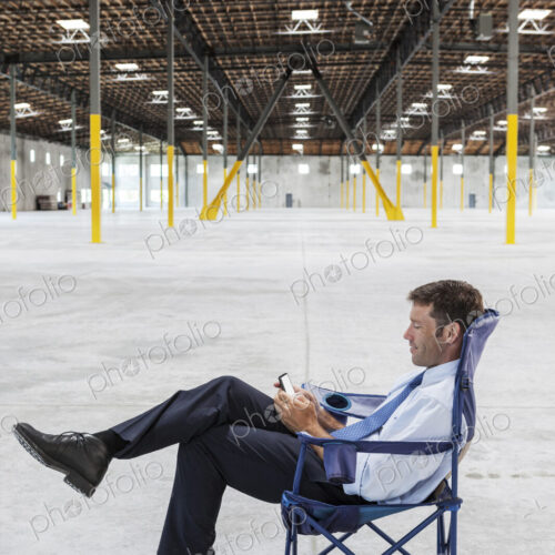 Caucasian man sitting in the middle of a new empty warehouse and using cell phone.