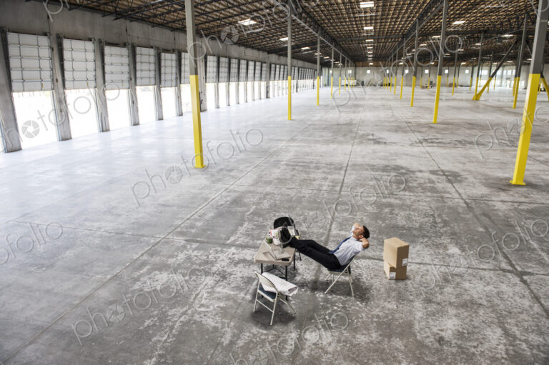Caucasian man sitting with feet up on desk in front of loading dock doors in a new…