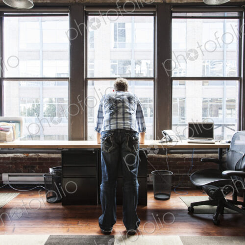 Caucasian man standing near a bank of windows at his creative office workstation.