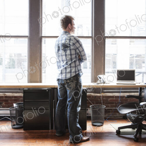 Caucasian man standing near a bank of windows at his creative office workstation.