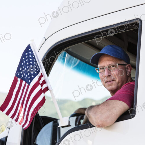 Caucasian man truck driver in the cab of his truck with an American Flag attached to… Caucasian man truck driver in the cab of his truck with an American Flag attached to…