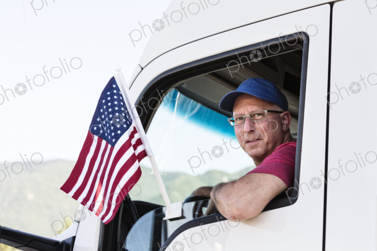 Caucasian man truck driver in the cab of his truck with an American Flag attached to…
