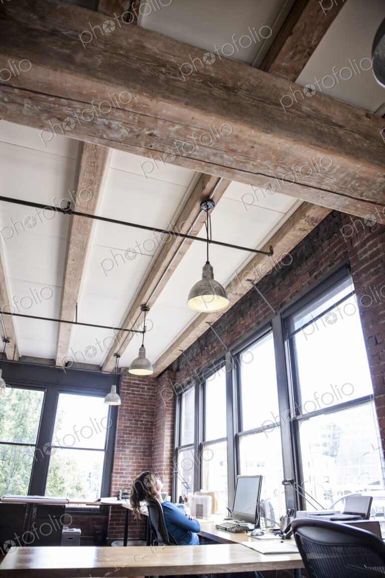 Caucasian woman at office workstation near a large bank of windows.