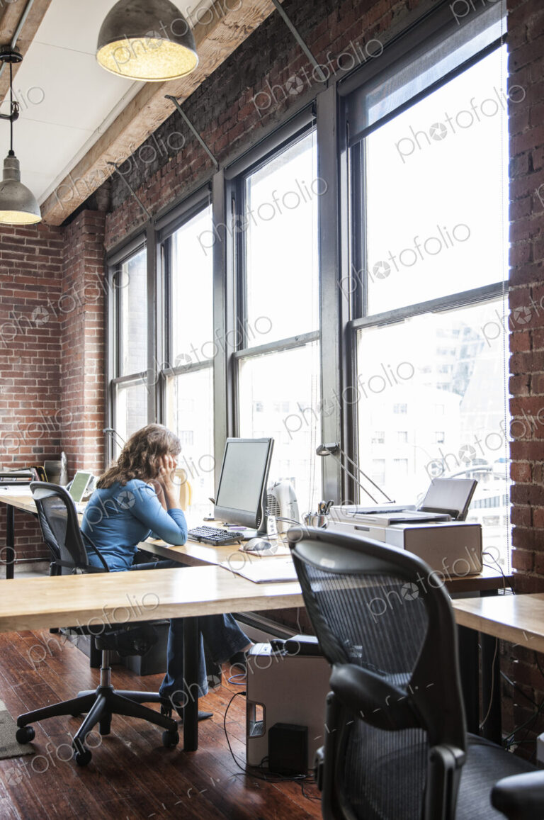 Caucasian woman at office workstation near a large bank of windows.