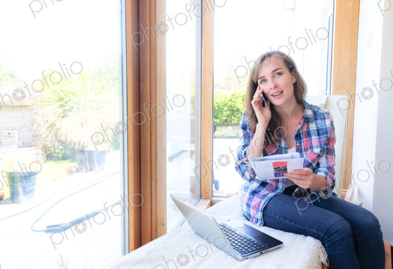 Caucasian woman holding paperwork and talking on cell phone near window