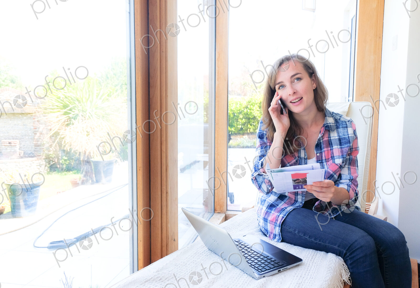 Caucasian woman holding paperwork and talking on cell phone near window