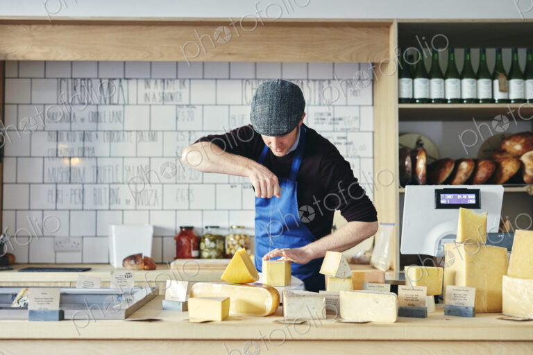 Cheesemonger cutting cheese with cheese wire surrounded by a variety of cheese on…