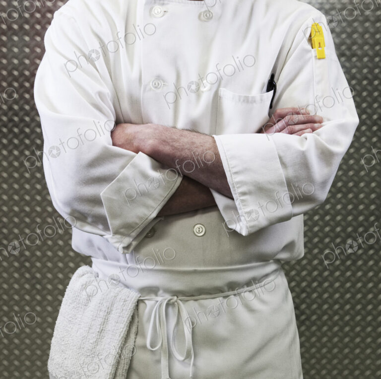 Chef in a white jacket and apron around his waist with arms folded. Mid section.