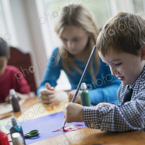 Children in a family home. Three children sitting at a table using glue and paint to…