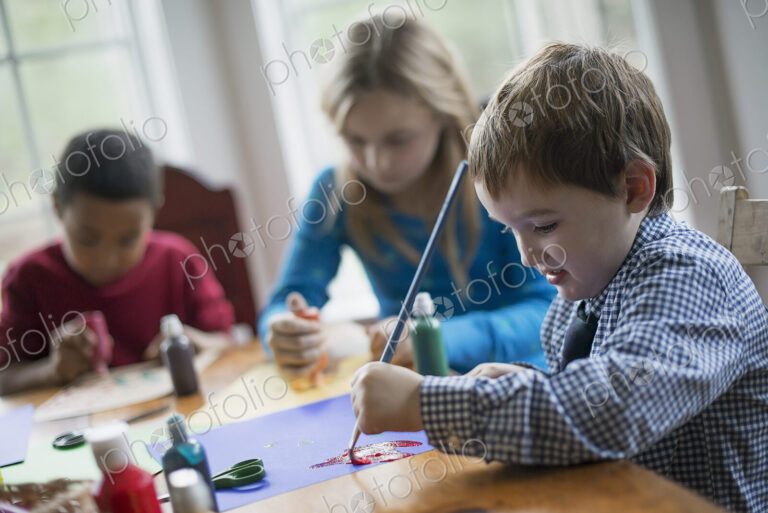 Children in a family home. Three children sitting at a table using glue and paint to…