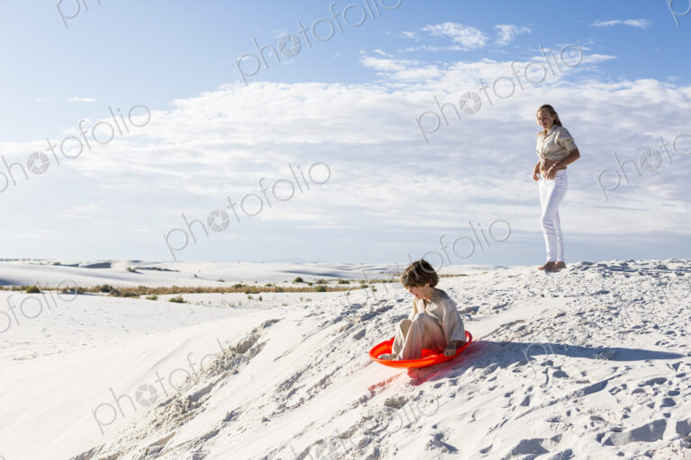 Children playing in sand dunes landscape, one on an orange sled.