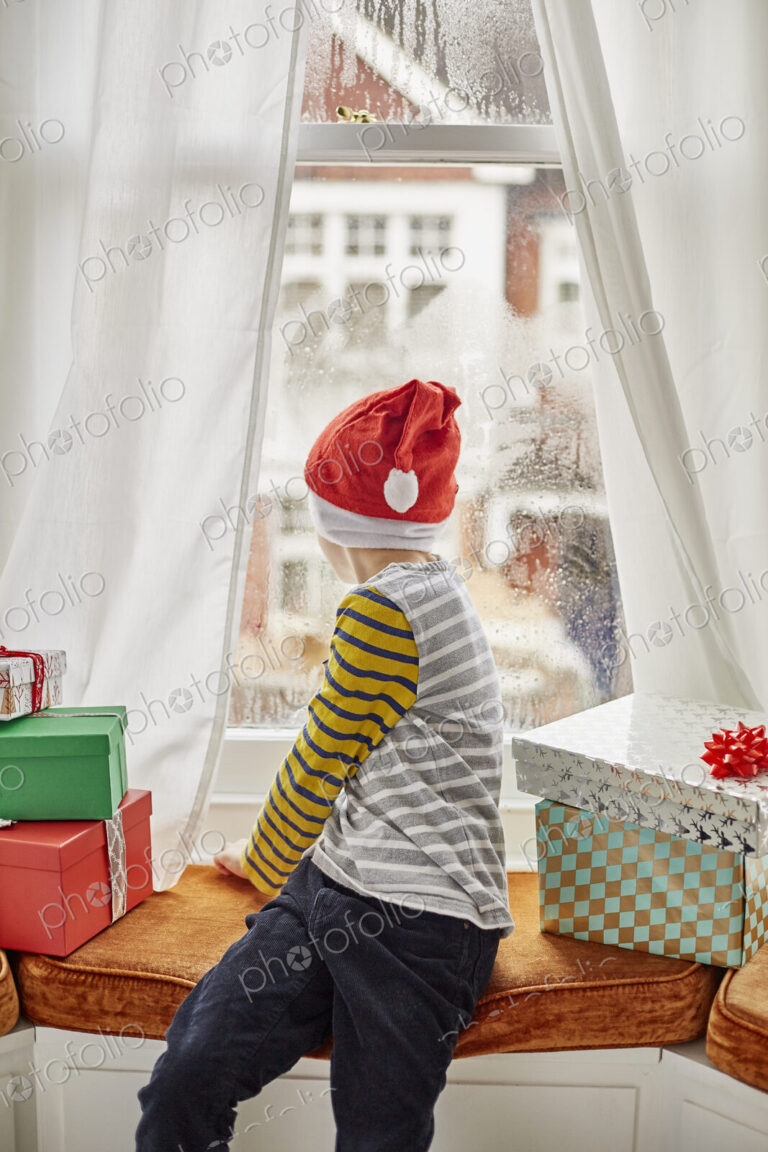 Christmas morning in a family home. A boy in a Santa hat looking out of a bedroom window.