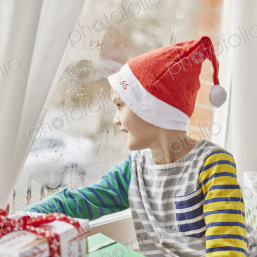 Christmas morning in a family home. A boy in a Santa hat looking out of a bedroom window. Christmas morning in a family home. A boy in a Santa hat looking out of a bedroom window.
