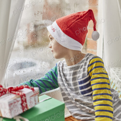 Christmas morning in a family home. A boy in a Santa hat looking out of a bedroom window. Christmas morning in a family home. A boy in a Santa hat looking out of a bedroom window.