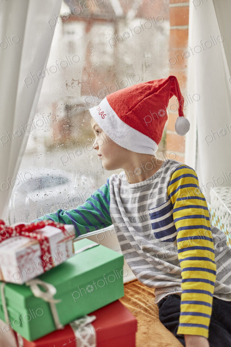 Christmas morning in a family home. A boy in a Santa hat looking out of a bedroom window.