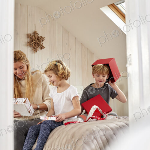 Christmas morning in a family home. A mother and two children sitting on a bed opening… Christmas morning in a family home. A mother and two children sitting on a bed opening…