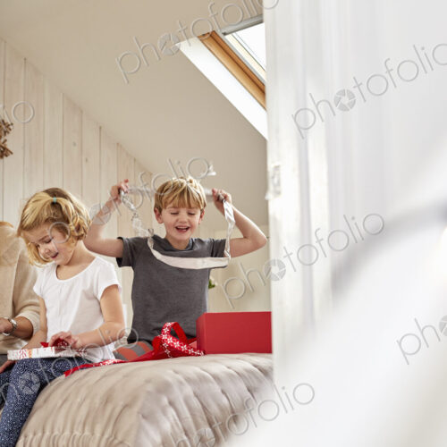 Christmas morning in a family home. A mother and two children sitting on a bed opening… Christmas morning in a family home. A mother and two children sitting on a bed opening…