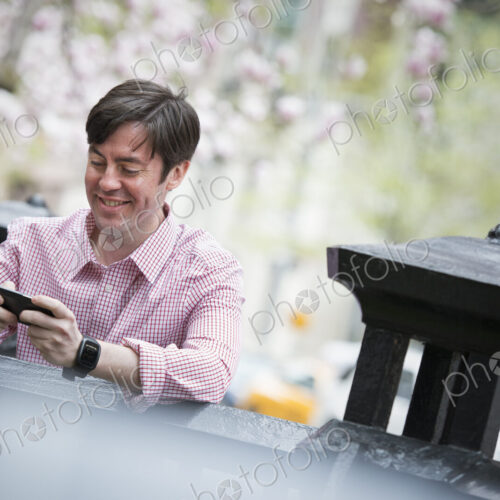 City life in spring. A man sitting outdoors in a city park. Looking down at his smart…