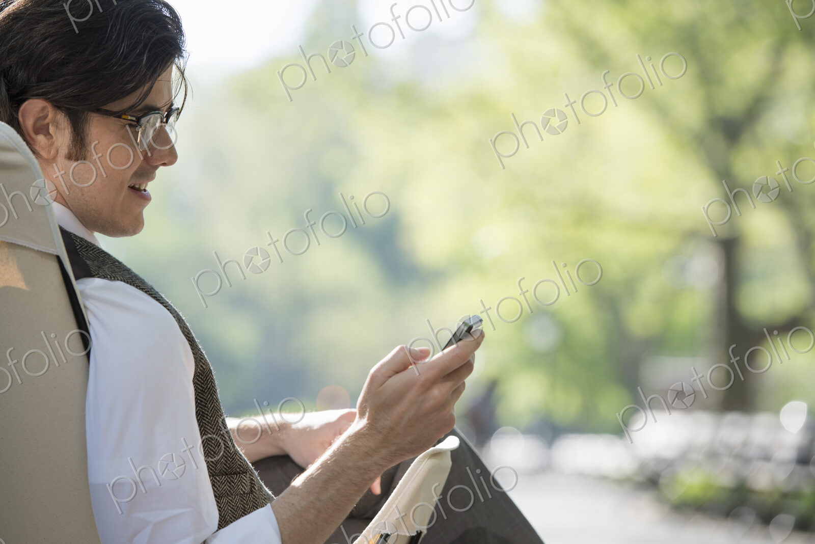 City life. A man sitting in a canvas camping chair in the park looking at his cell phone.