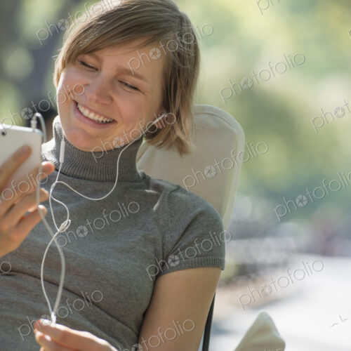 City life. A woman sitting in a camping chair in the park, listening to music wearing…