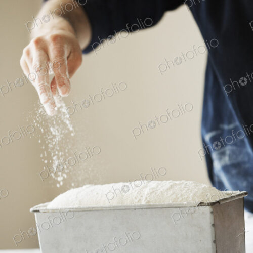 Close up of a baker sprinkling flour over bread dough in a metal baking tin.