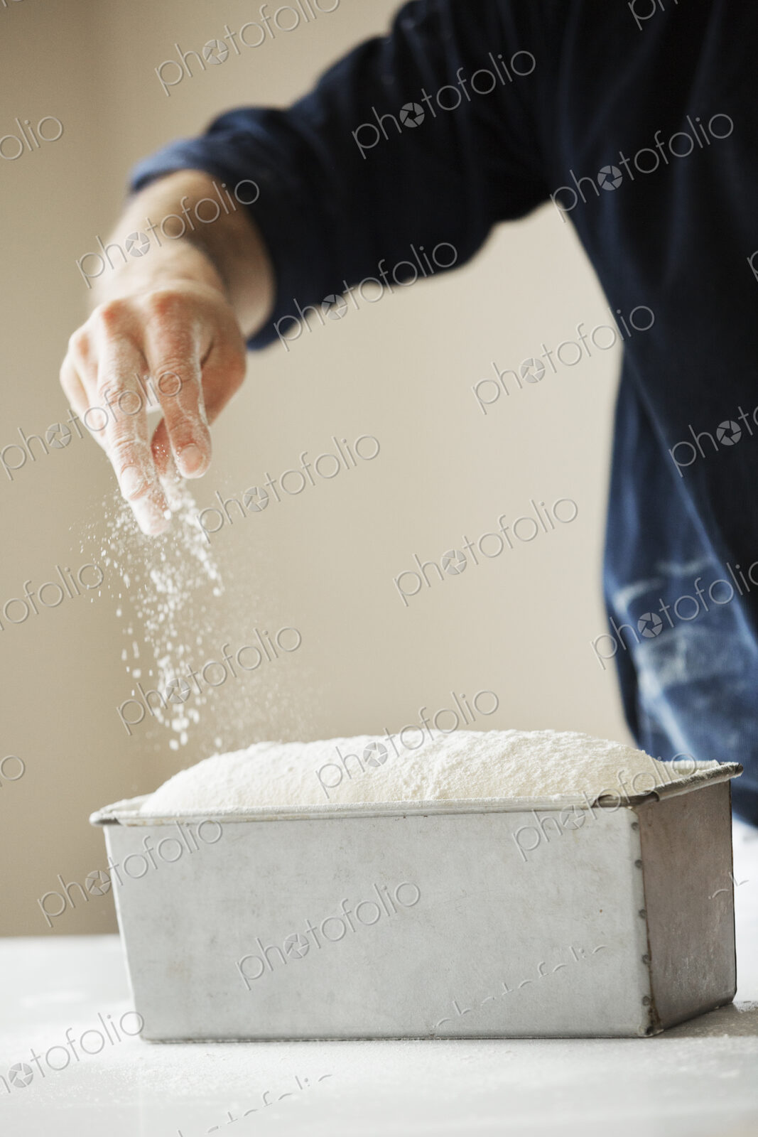 Close up of a baker sprinkling flour over bread dough in a metal baking tin.