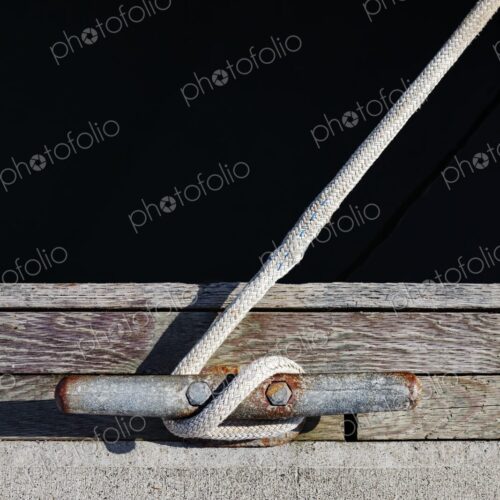 Close-up of a braided rope tied to a weathered and rusted dock cleat, set against aged…