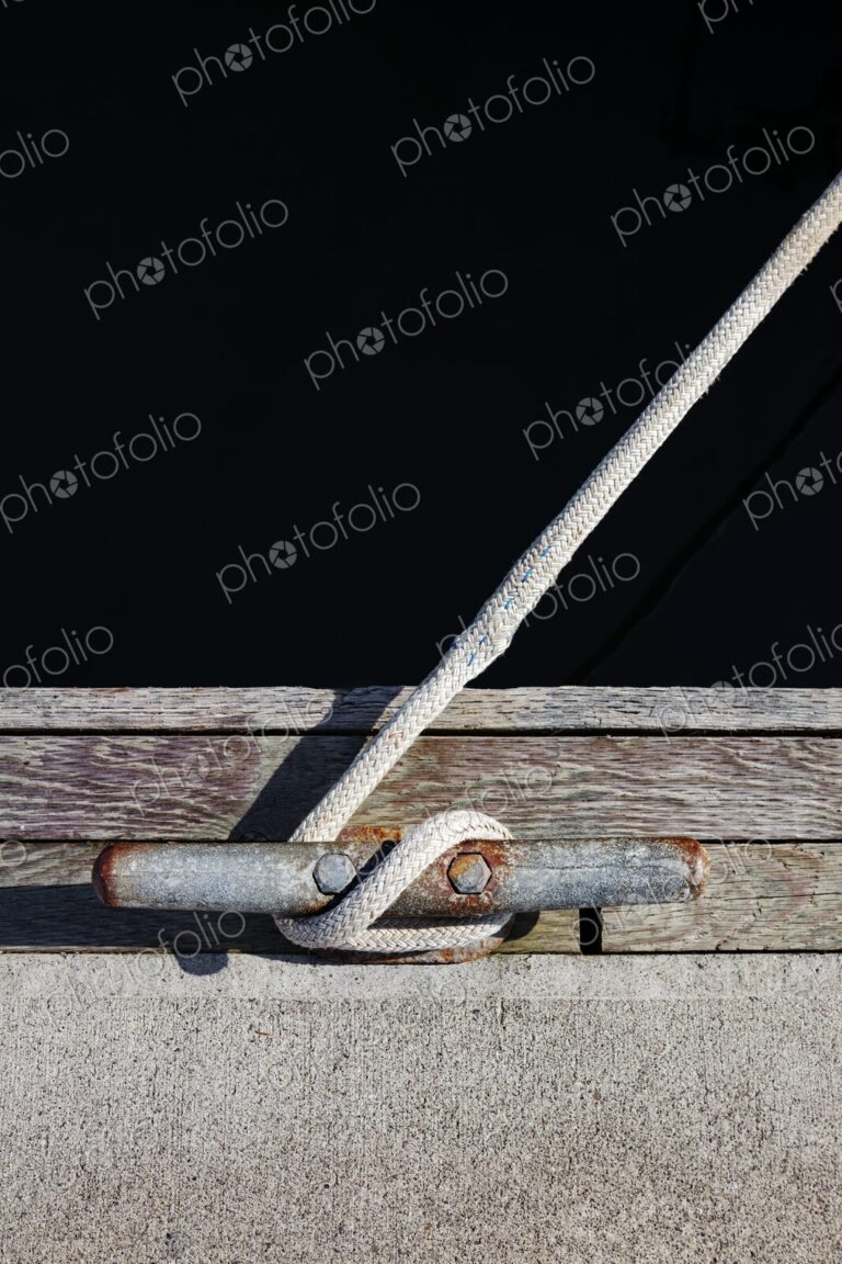 Close-up of a braided rope tied to a weathered and rusted dock cleat, set against aged…