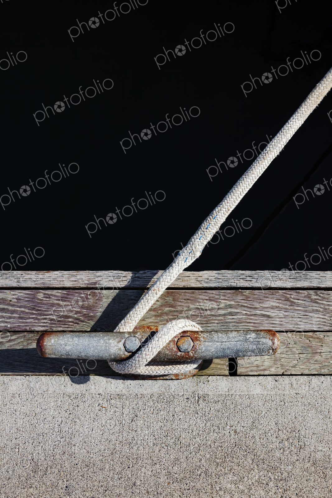 Close-up of a braided rope tied to a weathered and rusted dock cleat, set against aged…