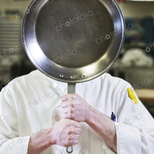 Close-up of a chef holding a sauté pan in front of his face in a kitchen. Close-up of a chef holding a sauté pan in front of his face in a kitchen.