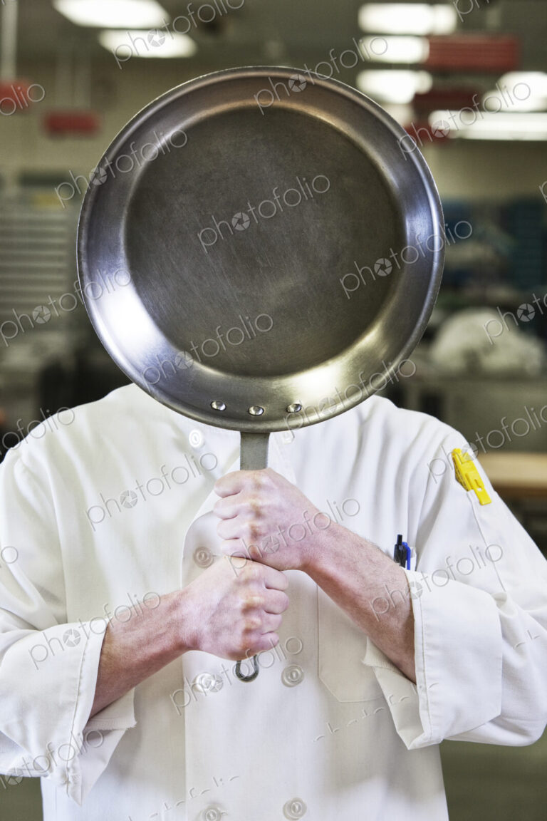 Close-up of a chef holding a sauté pan in front of his face in a kitchen.