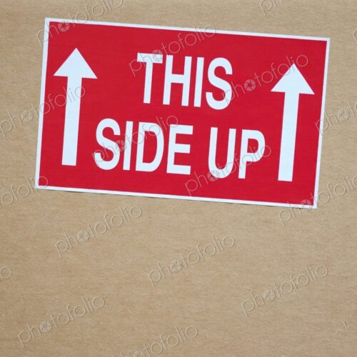 Close-up of a red "This Side Up" sticker with white arrows on a cardboard shipping box…