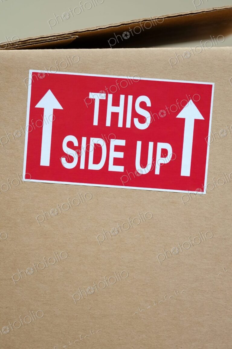 Close-up of a red "This Side Up" sticker with white arrows on a cardboard shipping box…