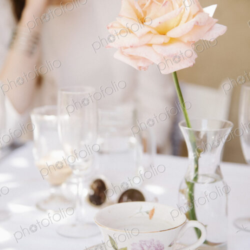 Close up of a rose in a vase on a table with a cup and saucer and glasses.