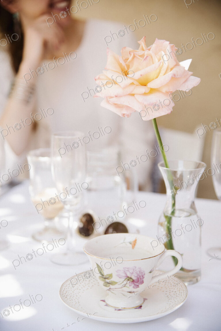 Close up of a rose in a vase on a table with a cup and saucer and glasses.