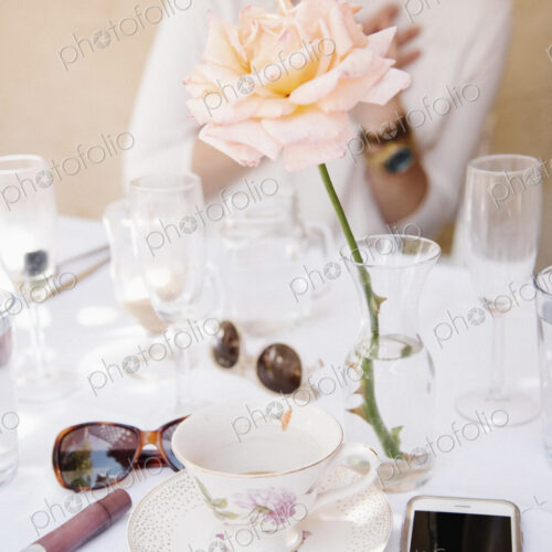 Close up of a rose in a vase on a table with cups and glasses, a cell phone and…