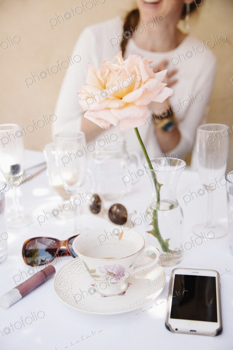 Close up of a rose in a vase on a table with cups and glasses, a cell phone and…